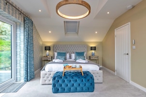 Un dormitorio con una cama grande, un reposapiés capitonado, una lámpara de techo circular y vistas a través de una ventana con cortinas.