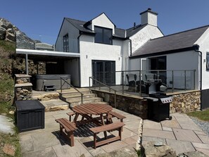A modern house with a patio area featuring a wooden table and benches, a barbecue grill, and a hot tub. - Stunning Bude Retreat With Hot Tub (Holyhead)