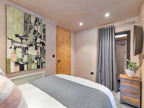A bedroom with a bed, a large abstract painting, a wooden door, a grey curtain, and a small plant on a wooden cabinet. - Selsey Retreat Near the Beach (Chichester)
