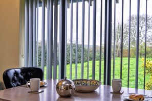 A dining table with a teapot, cups, and a plate with food, set against a window with vertical blinds. - Scenic Hot Tub Cottage for 2 (Wimborne)
