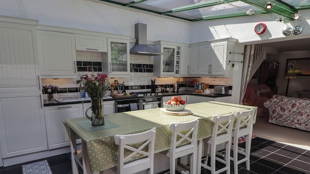 A kitchen with white cabinets, a dining table with a green tablecloth, and a vase with flowers. - The Garden Rooms (Narberth)
