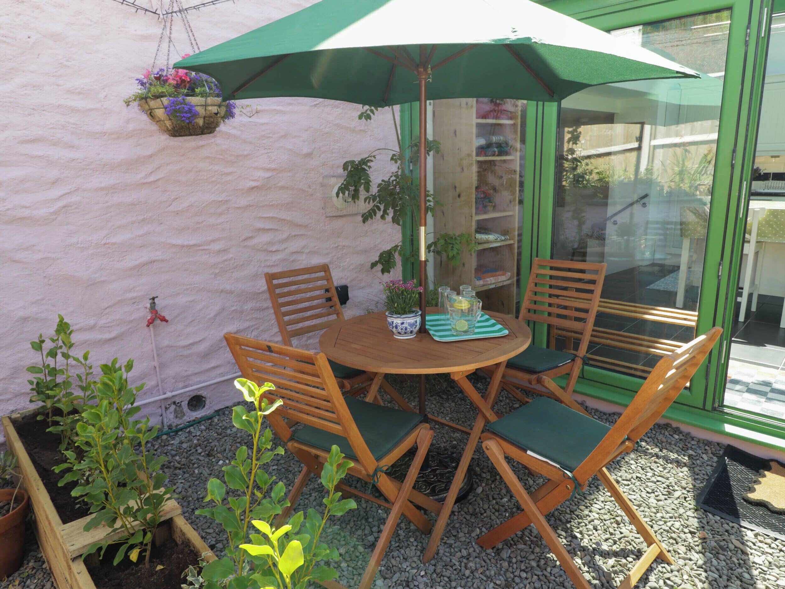 A round wooden table with four chairs, a hanging flower basket, and a green umbrella providing shade.