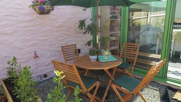 A round wooden table with four chairs, a hanging flower basket, and a green umbrella providing shade.