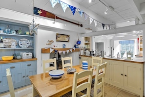 A kitchen with a wooden table, chairs, and various kitchen appliances. There are flags hanging from the ceiling, and a window with a view of the sea. - Estuary View Fowey Cottage for 6 (Fowey)