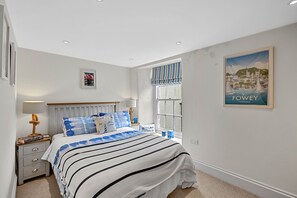 A bedroom with a bed, bedside tables, a window with curtains, a poster on the wall, and a lamp. - Estuary View Fowey Cottage for 6 (Fowey)