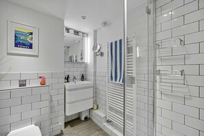 A modern bathroom with white tiled walls, a glass shower enclosure, a white sink with a mirror above it, and a framed picture on the wall. - Estuary View Fowey Cottage for 6 (Fowey)