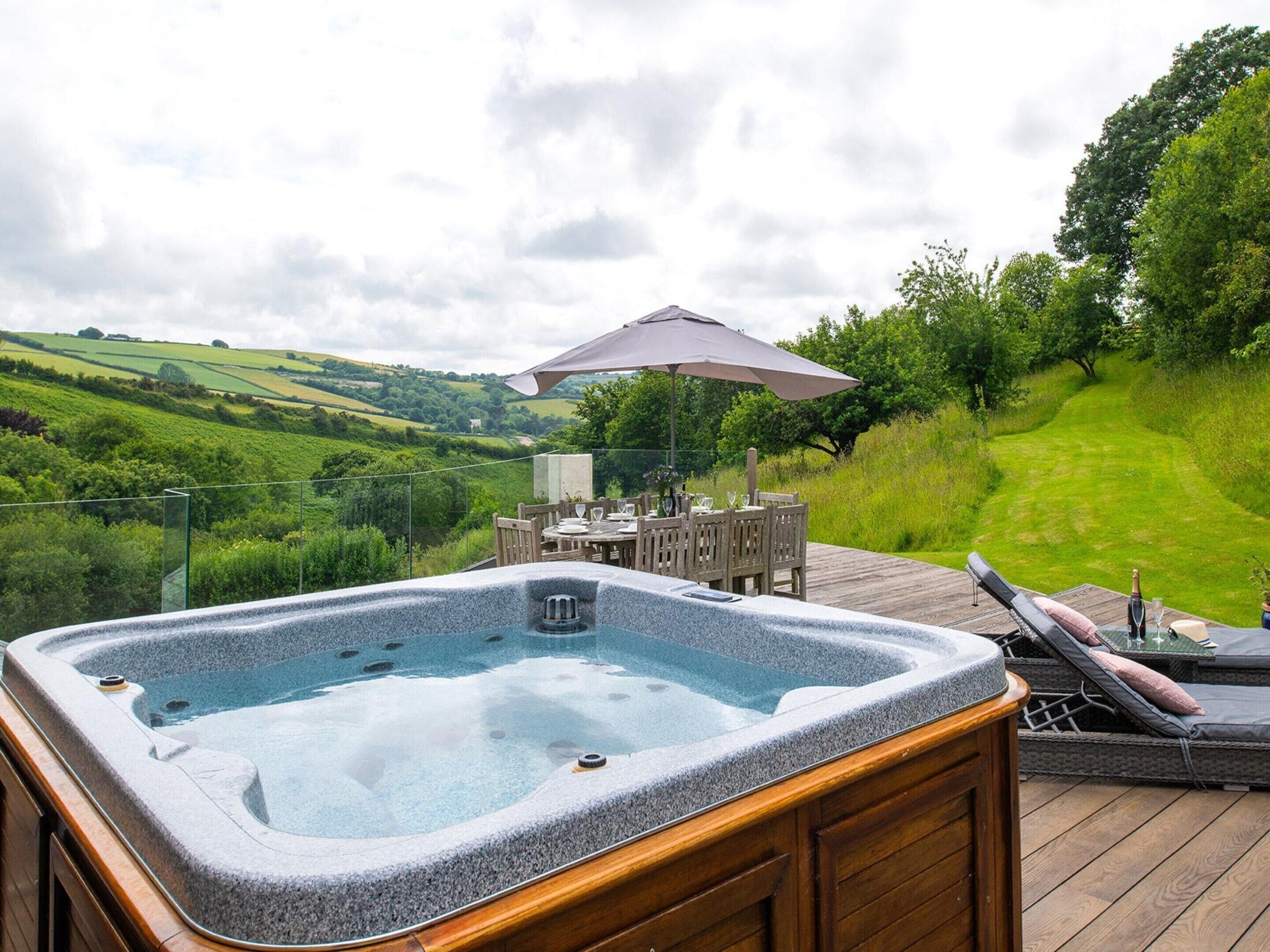 A hot tub on a wooden deck with a table and chairs, overlooking a green landscape.
