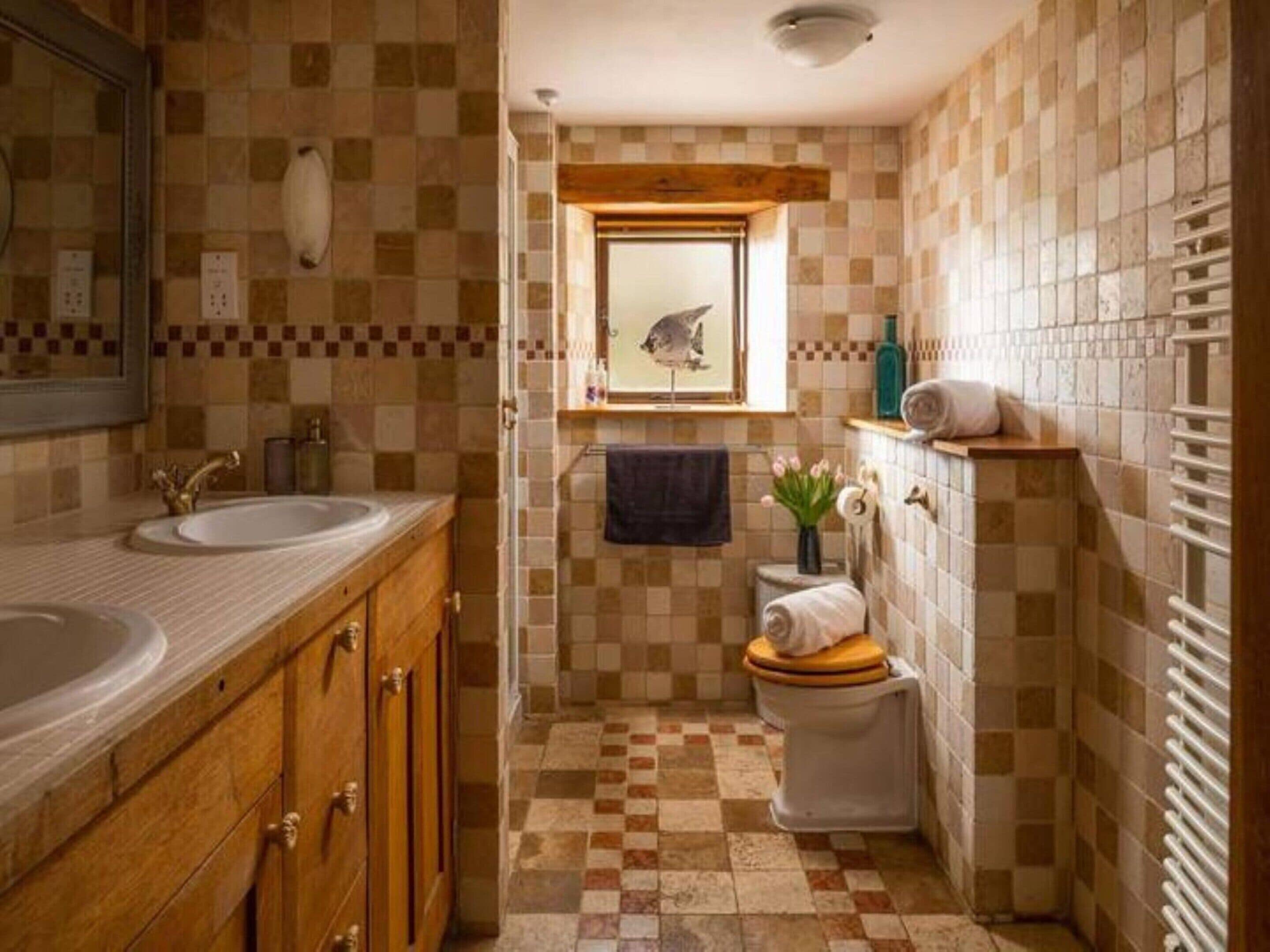 Un baño con piso a cuadros, un tocador de madera con lavamanos, un inodoro y una ventana con una toalla colgada en la pared.