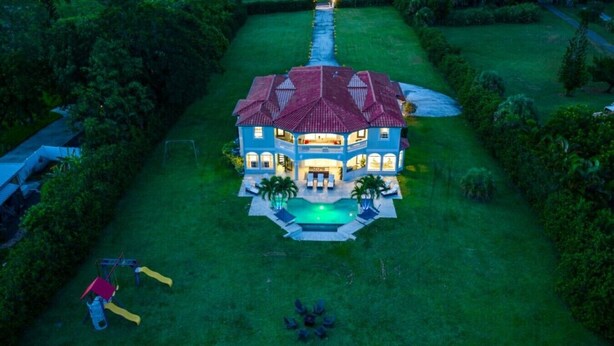 A large house with a swimming pool, surrounded by a well-maintained lawn and trees. - Riverfront Estate | Pool, Dock & Game Room (Palm City)