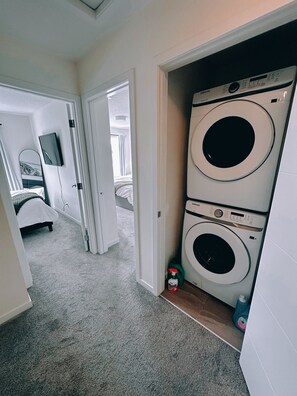 A laundry room with two stacked washing machines, a door leading to a bedroom, and a television mounted on the wall. - Fully equipped townhouse with a garage, 14 minutes from YYC Airport. Wi-Fi. (Airdrie)