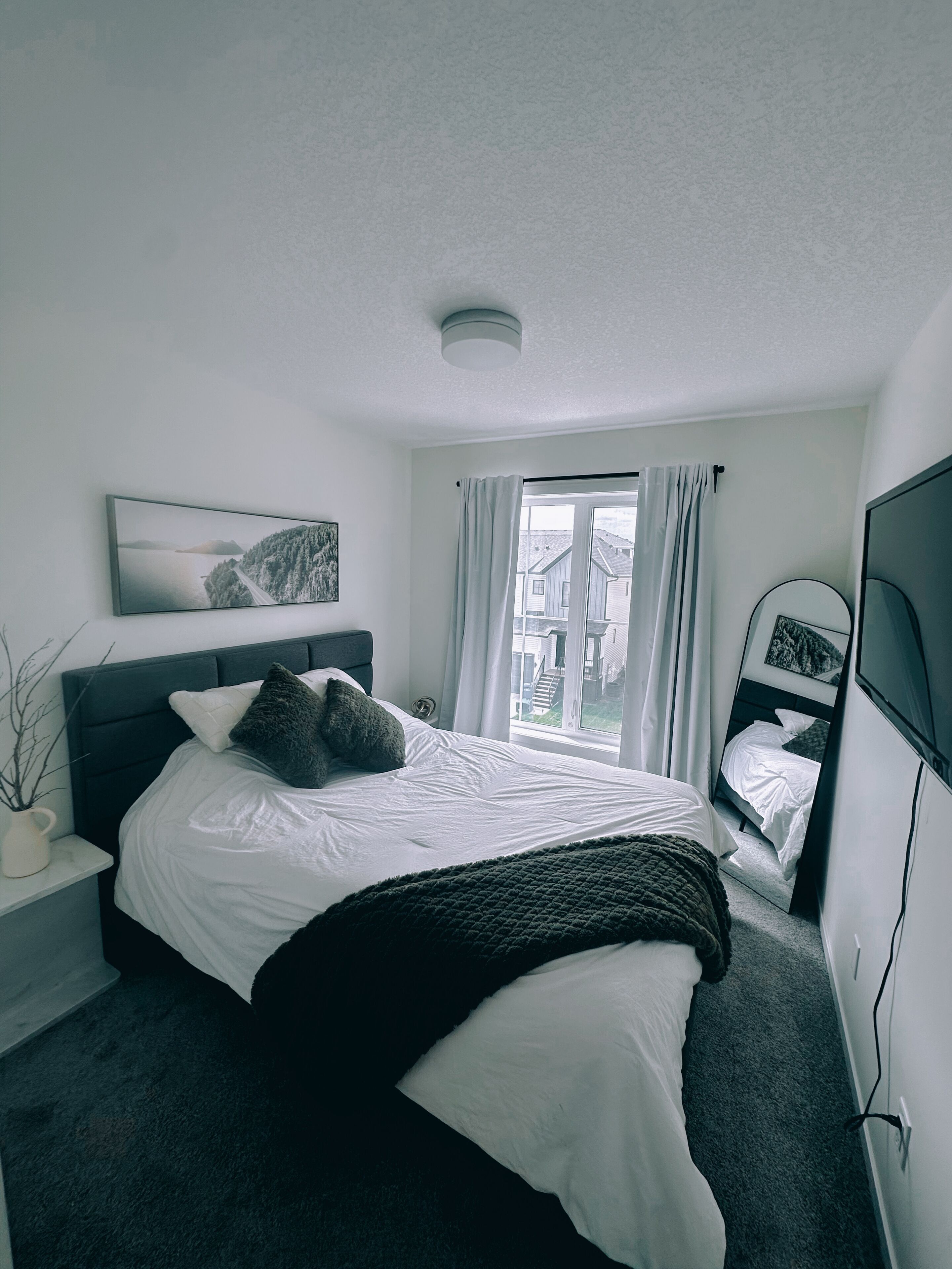 A bedroom with a bed, a bedside table, a window with curtains, a wall-mounted TV, and a framed picture.