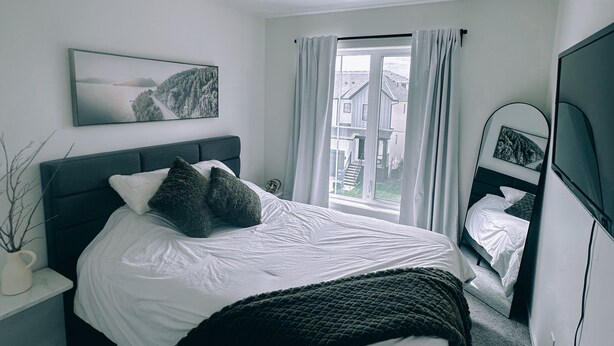 Un dormitorio con cama, mesita de noche, ventana con cortinas, televisor montado en la pared y un cuadro enmarcado.