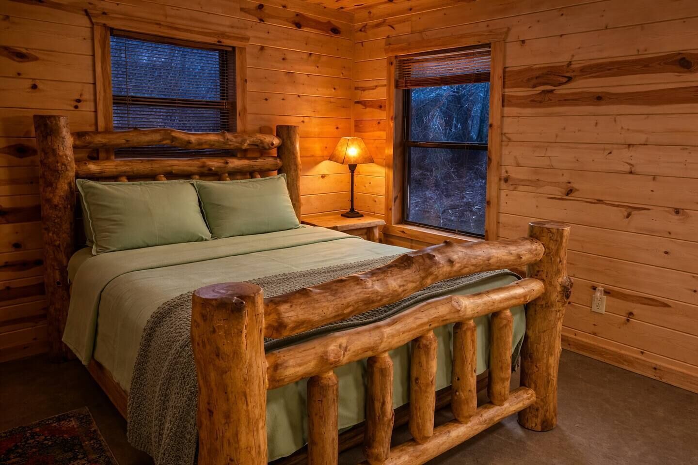 Un dormitorio de cabaña de madera con cama, almohadas, mesita de noche con lámpara y ventanas.