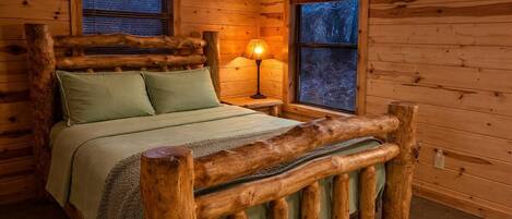 Un dormitorio de cabaña de madera con cama, almohadas, mesita de noche con lámpara y ventanas.