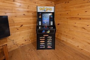 Ein Pac-Man-Spielautomat in einem Raum mit Holzwänden und einem Fernseher links.