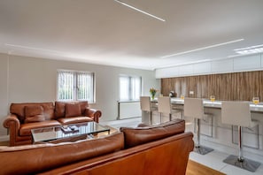 Una sala de estar moderna con un sofá de cuero marrón, una mesa de centro de vidrio y una zona de cocina con taburetes de bar.