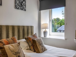 A hotel room with a bed, a window with a view of greenery, a lamp, and a wall with a rose pattern. - Barn Cottage With Hot Tub (Colintraive)