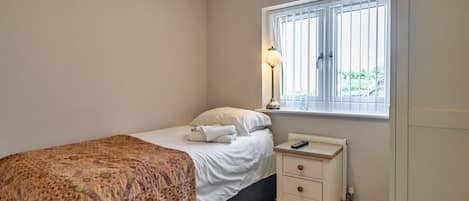 A bedroom with a bed, a bedside table with a lamp, a window with blinds, and a radiator.