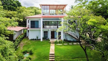 A modern two-story house with a large balcony, surrounded by lush greenery.