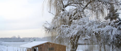A wooden cabin with a snow-covered roof, surrounded by a fence and a tree with hanging branches.