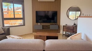 A living room with a flat-screen TV, wooden furniture, a round mirror, and a view of the outside. - AmdarVillage Cozy Garden Apt. (Eilat)