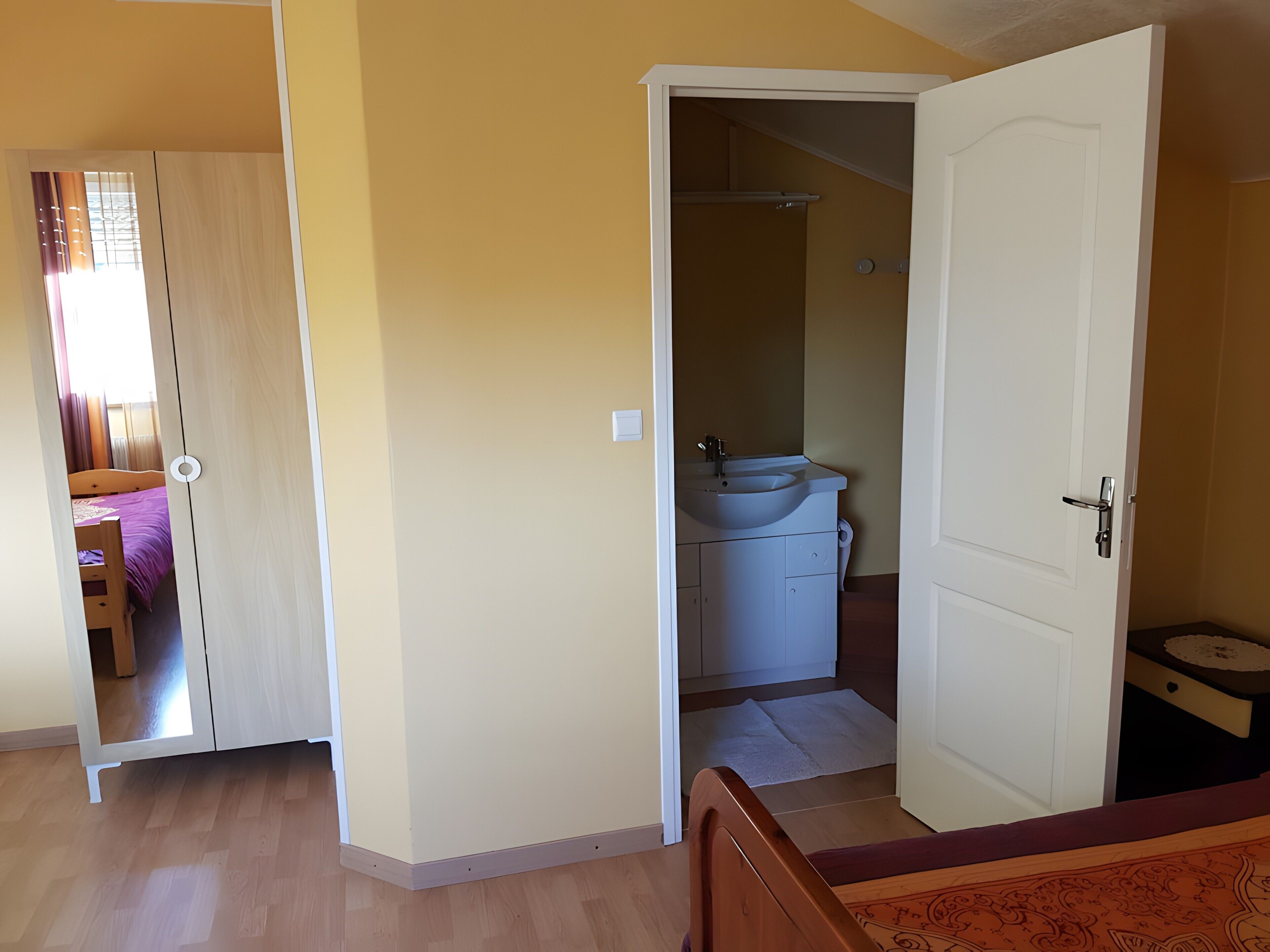 A room with a wooden bed, a white wardrobe, and a bathroom with a sink and mirror.