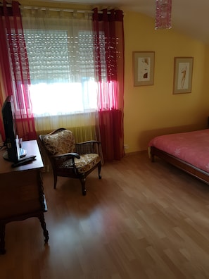 Una habitación con una cama, un escritorio con computadora, una silla y un ventanal con cortinas.