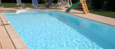 A rectangular swimming pool with a clear blue water surface, surrounded by a tiled deck. There are lounge chairs and a slide on the deck.