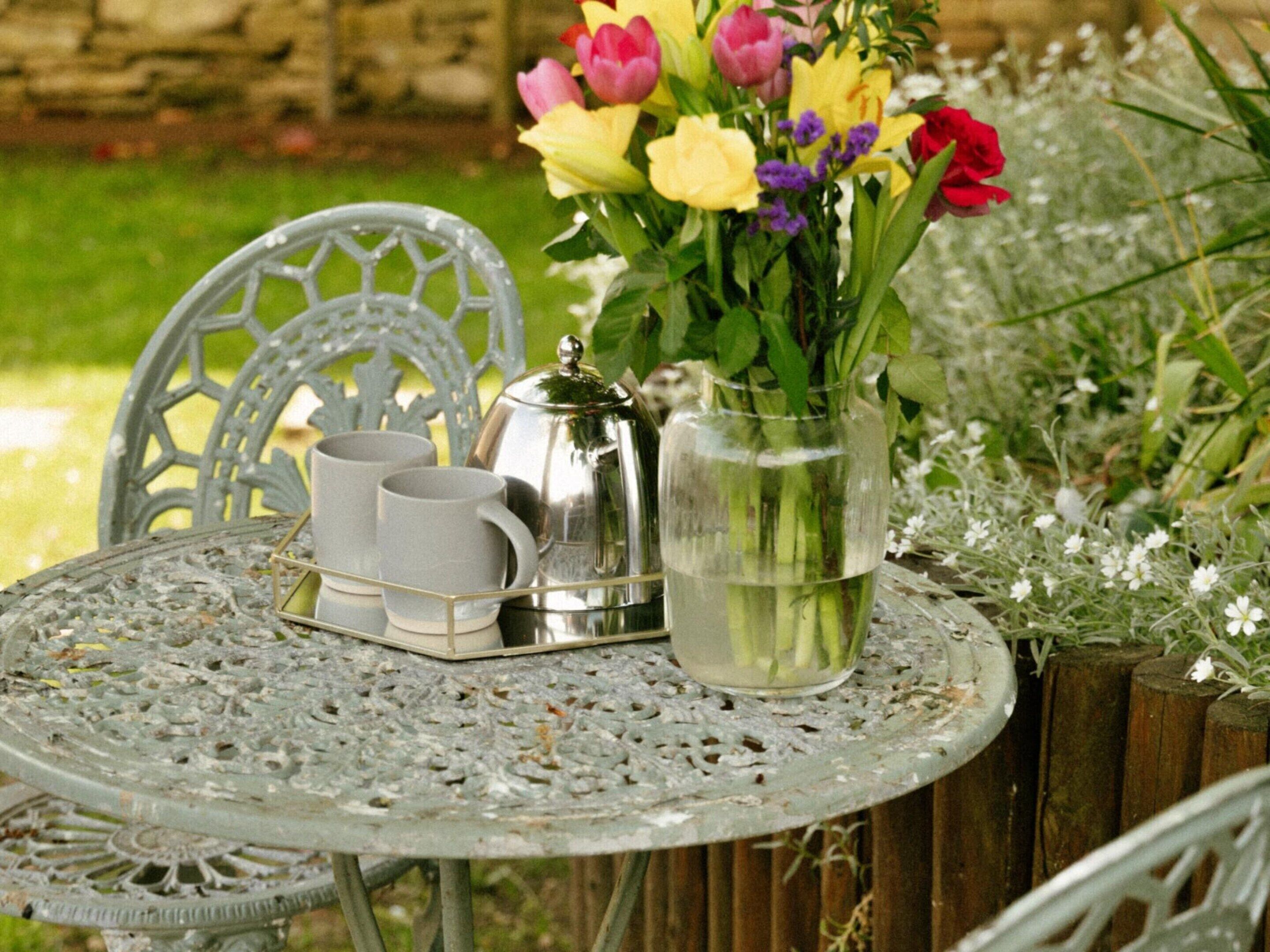 A metal table with a tray holding two cups and a silver pot, surrounded by chairs and a vase with flowers.