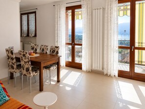 A dining area with a wooden table and patterned chairs, a small round table, and a view through a large window with curtains. - Stunning Pool Apartment With Lake View (Brezzo di Bedero)