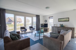 Ein modernes Wohnzimmer mit einer Couch, Sesseln, einem Couchtisch und einem Fernseher. Ein Balkon bietet Blick auf Gebäude und Bäume.