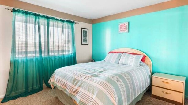A bedroom with a bed, a nightstand, a window with sheer curtains, and a wall with a framed picture. - Cozy 1-bedroom condo in delightful Ocean City Jockey Beach Club with AC (Ocean City)