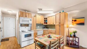A compact kitchen with wooden cabinets, a white stove, and a dining table with a colorful placemat. - Cozy 1-bedroom condo in delightful Ocean City Jockey Beach Club with AC (Ocean City)
