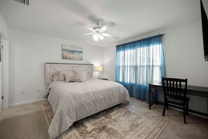 Ein Schlafzimmer mit einem Bett, einem Schreibtisch, einem Sessel, einem Deckenventilator und einem Fenster mit Vorhängen.