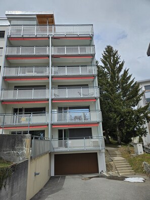 Un edificio residencial de varios pisos con balcones, un garaje y un árbol al fondo.