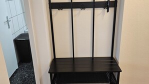 Un estante metálico negro con ganchos y un estante inferior, ubicado frente a una pared con suelo de baldosas.