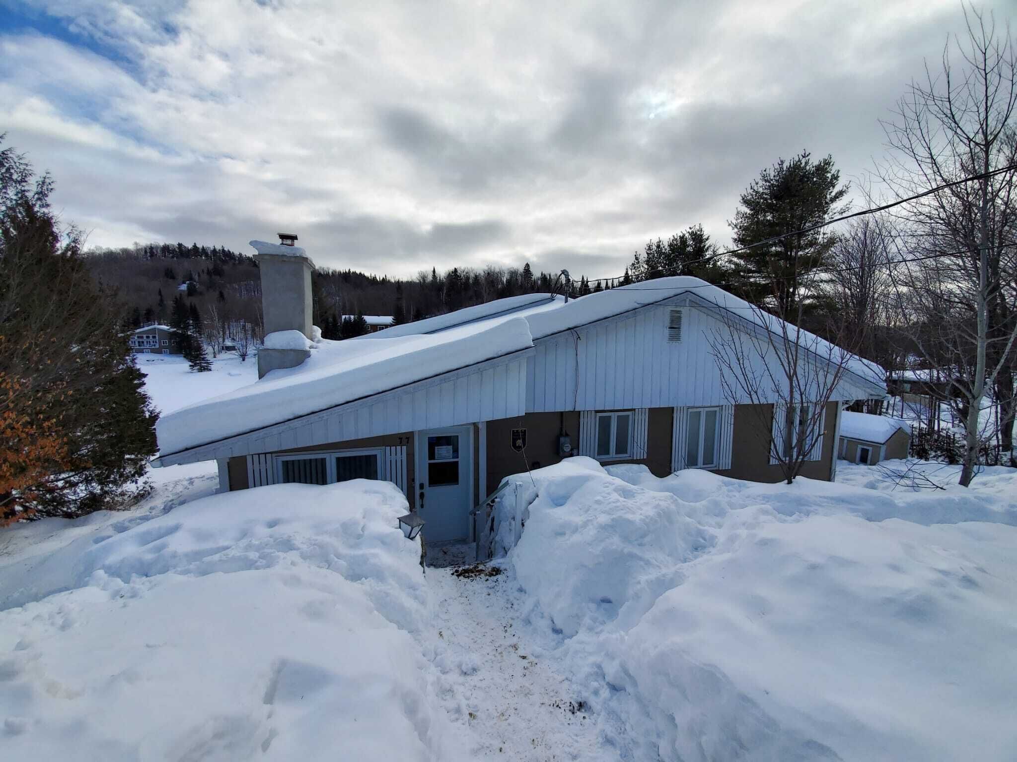 A snow-covered building with a metal roof, surrounded by snowbanks, and a forested backdrop.