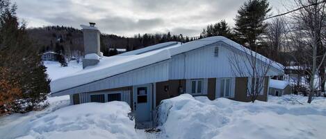 A snow-covered building with a metal roof, surrounded by snowbanks, and a forested backdrop.