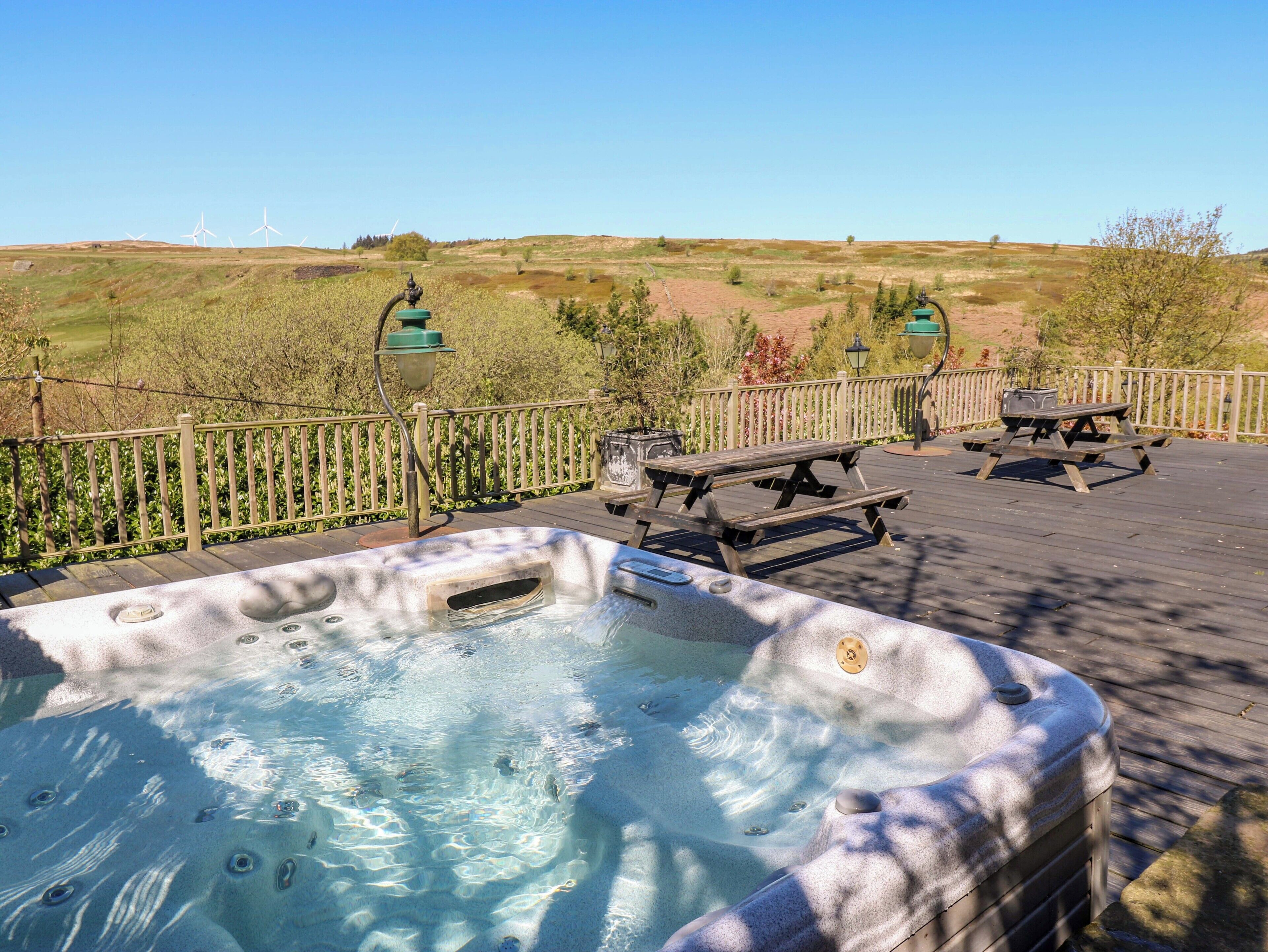Un jacuzzi en una terraza de madera con mesas de picnic y una cerca con vistas a un campo cubierto de hierba y aerogeneradores.