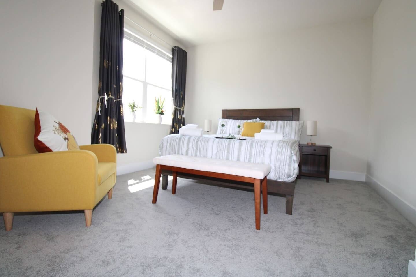 A bedroom with a bed, a bench, a nightstand, a lamp, and a window with curtains.