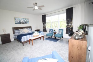 Een slaapkamer met een bed, twee stoelen, een bureau, een plafondventilator en een raam met gordijnen.