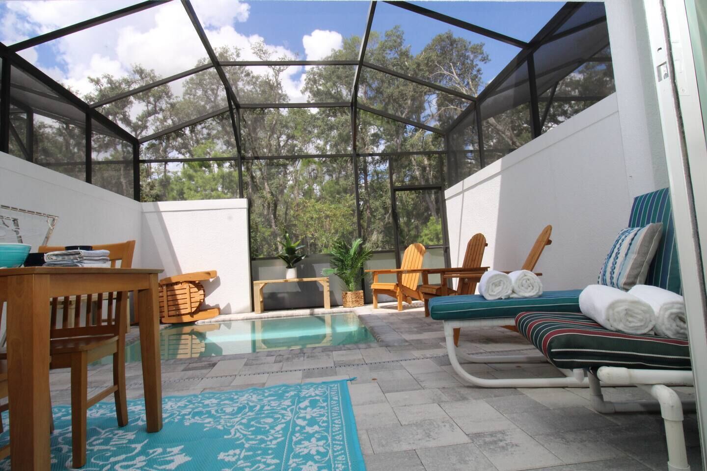 A sunroom with a glass ceiling, a pool, wooden furniture, and a lounge area with a view of trees.