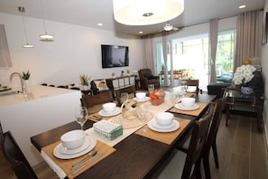 Een moderne eetruimte met een lange tafel voor zes personen, een keuken met een spoelbak en een televisie, en een woongedeelte met een bank en een salontafel.
