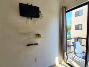 A room with a mounted TV, two white shelves, and a window with a view of buildings and greenery. - Dream 2-bedroom apartment in Puerto Morelos with pools, gym, tennis court & more (Puerto Morelos)