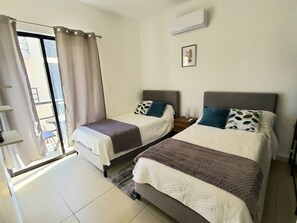 A hotel room with two beds, a window with curtains, an air conditioning unit, and a small picture on the wall. - Dream 2-bedroom apartment in Puerto Morelos with pools, gym, tennis court & more (Puerto Morelos)
