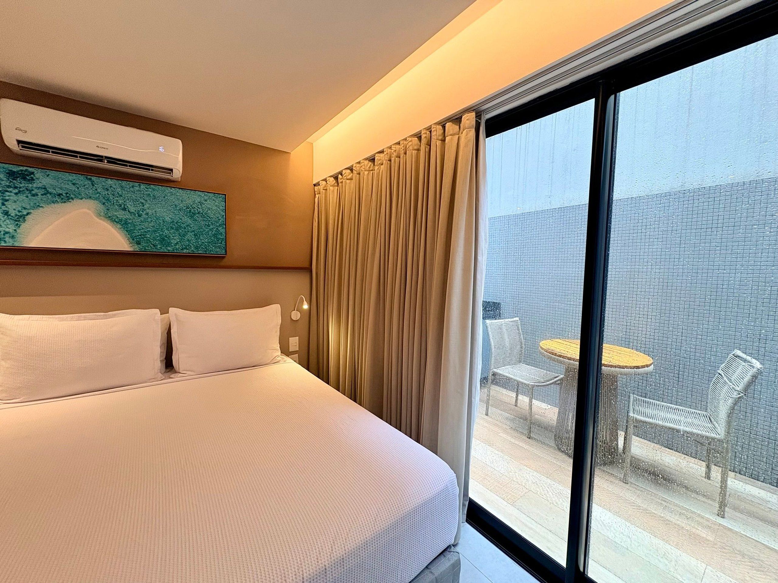 Une chambre d’hôtel dotée d’un grand lit, d’un appareil de climatisation, d’une œuvre d’art accrochée au mur et offrant une vue sur un espace extérieur aménagé, visible par une fenêtre.