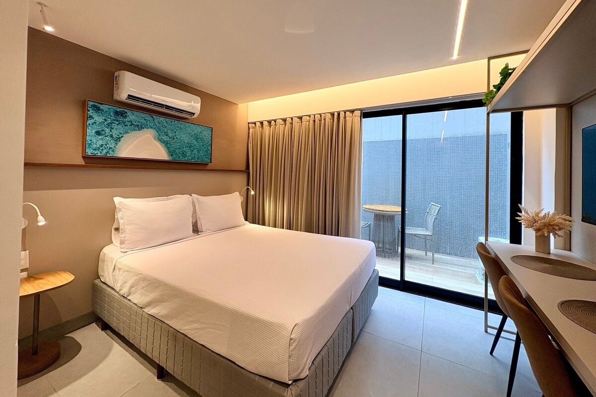 A modern hotel room with a large bed, a desk, and a view of the cityscape through the balcony.