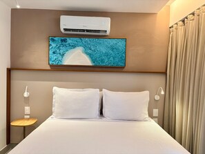 Una camera d'albergo con un letto, un condizionatore d'aria a parete, una TV fissata al muro, un comodino con una lampada e una finestra con tende.