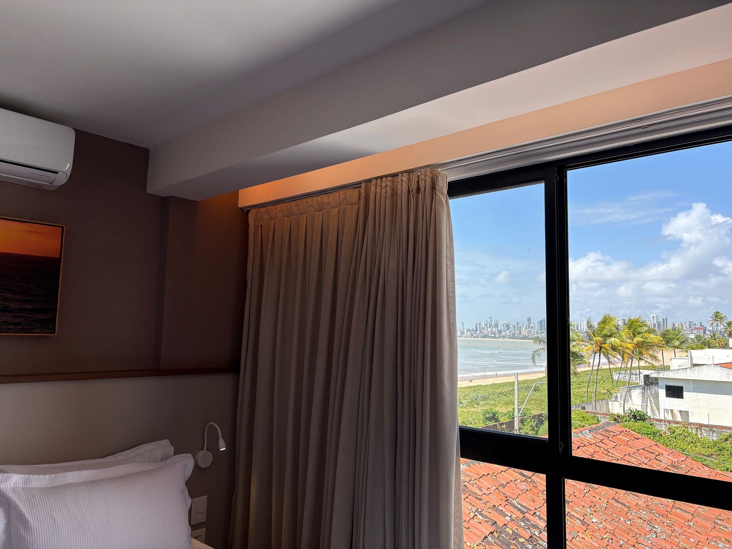 A hotel room with a large window, a bed, an air conditioning unit, and a view of a cityscape and beach.
