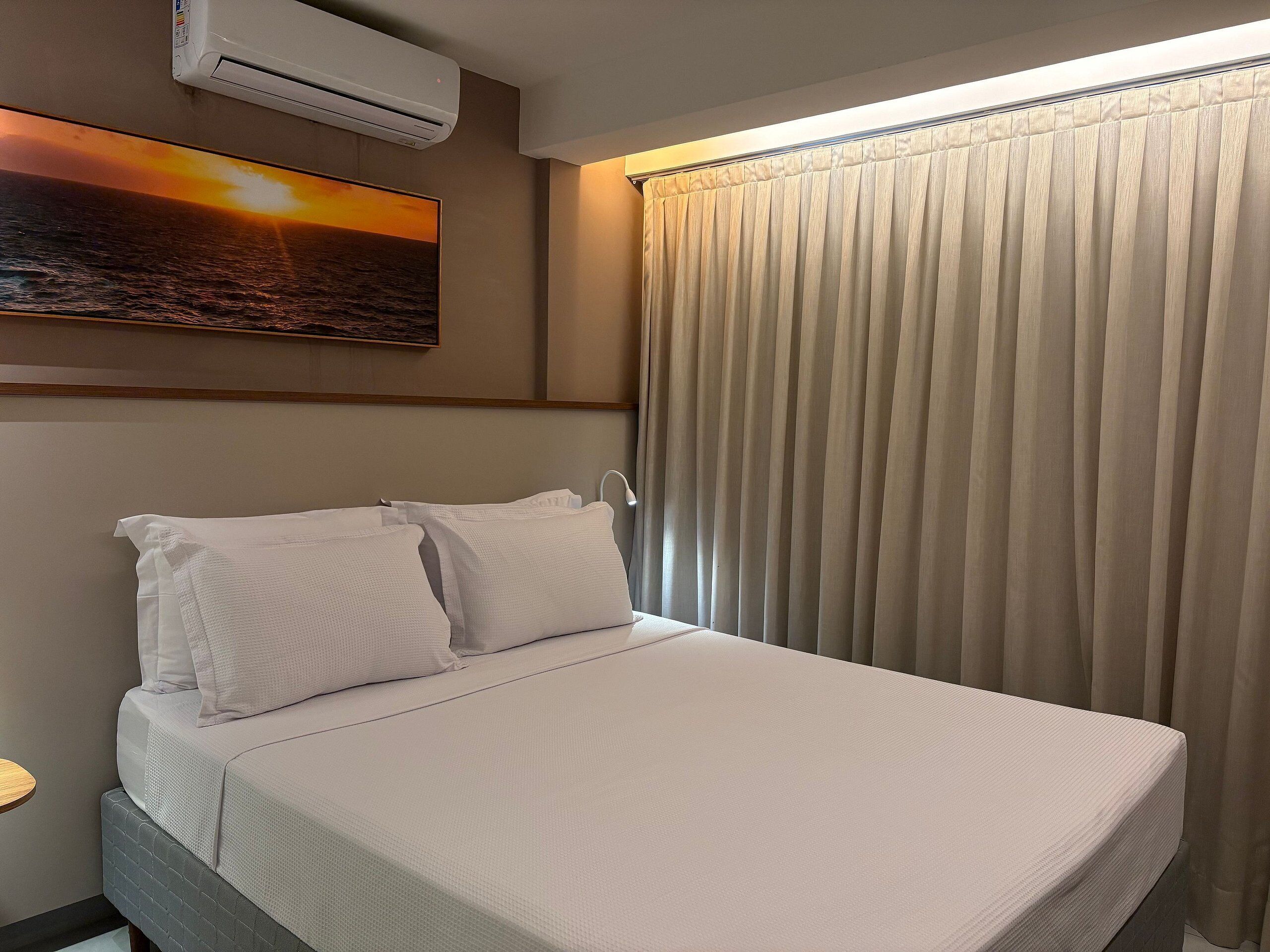 A hotel room with a bed, a wall-mounted air conditioner, a painting of a sunset, and a window with curtains.